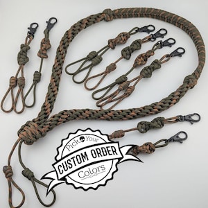 May include: A collection of olive green and brown paracord lanyards and attachments featuring black metal swivel clips. The braided cord items include various knot designs and loops. A white circular graphic with a skull icon displays the text Pick Your CUSTOM ORDER Colors.
