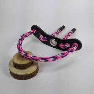 May include: A pink, white, and purple braided rope with a black leather loop and a metal ring. The rope is tied to a small wooden disc.