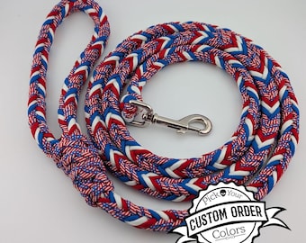 May include: A braided dog leash featuring a red, white, and blue pattern with a silver metal swivel snap hook. The leash includes a handle loop and a circular coiled section. A black and white graphic label reads Pick Your CUSTOM ORDER Colors.