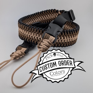 May include: A braided camera strap in shades of black, brown, and tan, featuring a black plastic buckle and tan cords. The strap is designed with a woven pattern. The image includes a logo that says "Pick Your Custom Order Colors."