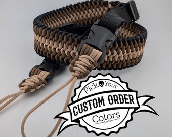 Könnte beinhalten: Ein geflochtener Kameragurt in Schwarz, Braun und Beige, mit einer schwarzen Kunststoffschnalle und beigefarbenen Kordeln. Der Gurt hat ein gewebtes Muster. Das Bild enthält ein Logo mit der Aufschrift "Pick Your Custom Order Colors."
