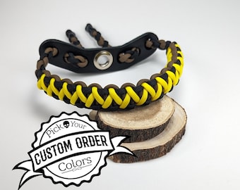 May include: A black, brown, and yellow archery wrist sling. The sling is made of woven paracord and features a black leather pad with a metal grommet. The text "CUSTOM ORDER COLORS" is in a circular badge.