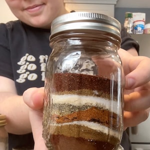 May include: A glass jar filled with layers of different spices, including chilli powder, cumin, oregano, and garlic powder. The jar is labelled "Made in USA".