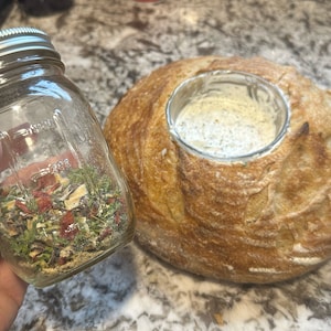 May include: A glass jar filled with dried herbs and spices, next to a round loaf of bread with a hollowed-out centre.