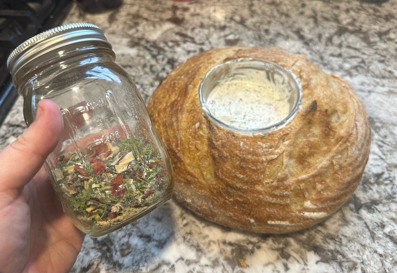 May include: A glass jar filled with dried herbs and spices, next to a round loaf of bread with a hollowed-out centre.