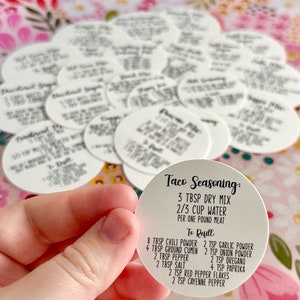 May include: White round labels with black text for taco seasoning. The label includes the ingredients and instructions for making taco seasoning. The text reads "Taco Seasoning: 3 tbsp dry mix 2/3 cup water per one pound meat To Refill: 8 tbsp chili powder 2 tsp garlic powder 4 tbsp ground cumin 2 tsp onion powder 2 tbsp pepper 2 tsp oregano 2 tbsp salt 4 tsp paprika 2 tsp red pepper flakes 2 tsp cayenne pepper"