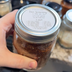 May include: A glass jar filled with brown taco seasoning mix. The lid has a white label with the text "Taco Seasoning: 3 Tbsp Dry Mix 2/3 Cup Water Per One Pound Meat To Reuft 4 Tbsp Chili Powder 2 Tbsp Ground Cumin 1 Tsp Garlic Powder 1 Tsp Onion Powder 1 Tbsp Salt 1 Tbsp Pepper 1 Tsp Oregano 2 Tsp Paprika 1 Tsp Red Pepper Flakes 1 Tsp Cayenne Pepper".
