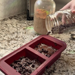 May include: A red silicone baking pan with two sections filled with chocolate brownie batter and topped with chocolate chips.