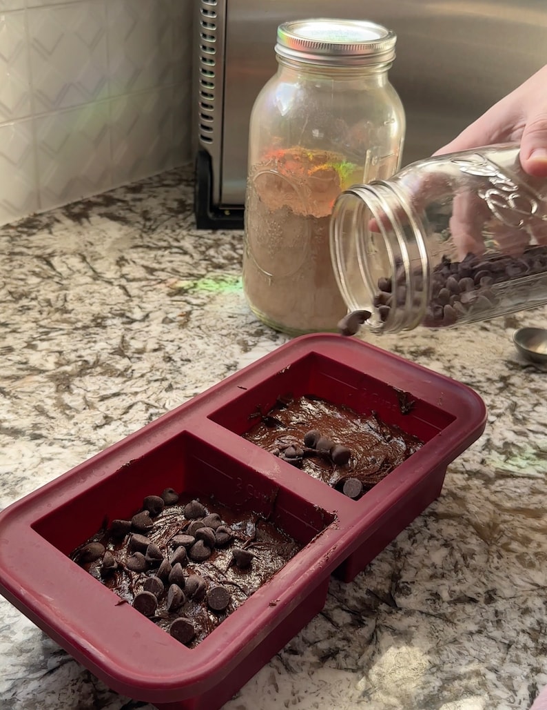 May include: A red silicone baking pan with two sections filled with chocolate brownie batter and topped with chocolate chips.