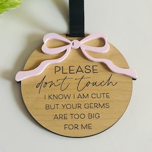 May include: A round wooden sign with the text "PLEASE don't touch" and the phrase "I KNOW I AM CUTE BUT YOUR GERMS ARE TOO BIG FOR ME". The sign has a pink bow and a black strap.