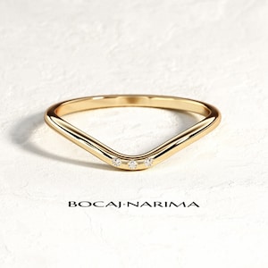 May include: A gold V-shaped ring with three small, clear gemstones set in the center. The ring is on a white surface, and the text "BOCAI•NARIMA" is printed below it.