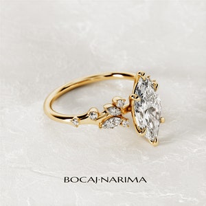 May include: A gold ring with a marquise-shaped diamond and a delicate floral design. The ring features a cluster of smaller diamonds set in a leaf-like pattern.