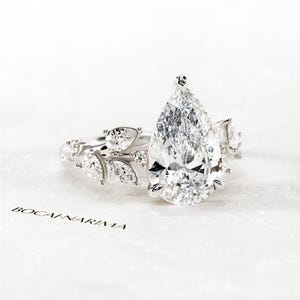 Puede incluir: Un anillo de compromiso de diamante de forma pera con un delicado dise&ntilde;o floral. El anillo presenta una gran piedra central de forma pera rodeada de diamantes redondos y de forma pera m&aacute;s peque&ntilde;os. La banda est&aacute; adornada con un delicado dise&ntilde;o floral, creando un look rom&aacute;ntico y elegante.