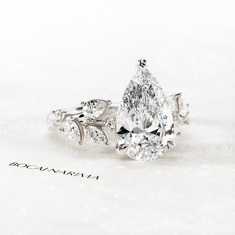 Puede incluir: Un anillo de compromiso de diamante de forma pera con un delicado dise&ntilde;o floral. El anillo presenta una gran piedra central de forma pera rodeada de diamantes redondos y de forma pera m&aacute;s peque&ntilde;os. La banda est&aacute; adornada con un delicado dise&ntilde;o floral, creando un look rom&aacute;ntico y elegante.