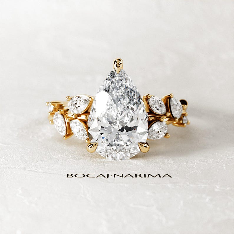 Puede incluir: Un anillo de compromiso de diamante de forma pera con una banda de oro. El anillo presenta un gran diamante de forma pera en el centro, rodeado de diamantes de forma pera m&aacute;s peque&ntilde;os. El anillo est&aacute; engastado en un delicado dise&ntilde;o floral.