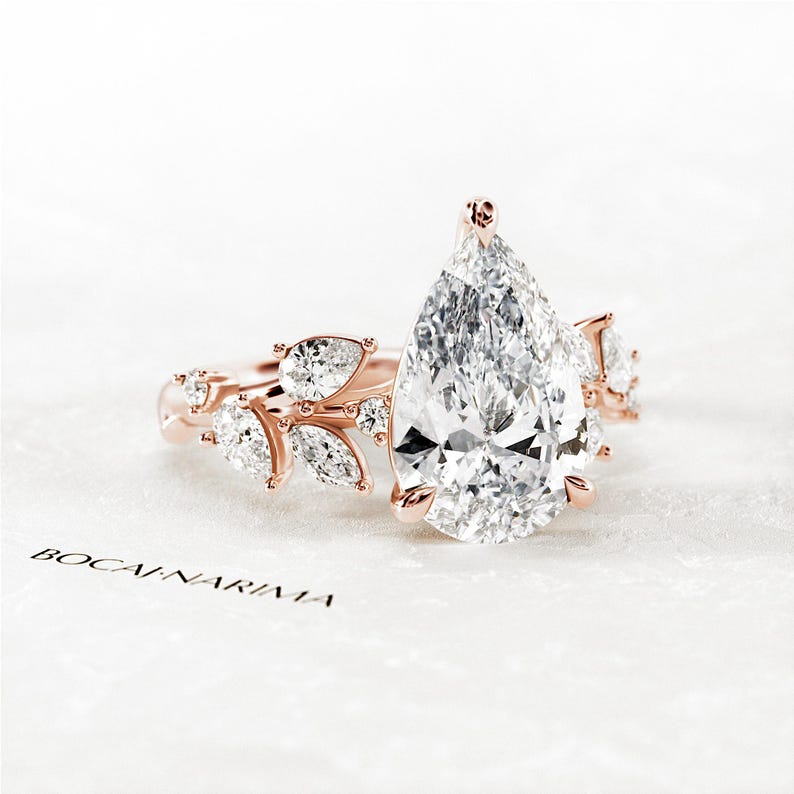 Puede incluir: Un anillo de compromiso de diamante de forma pera engastado en oro rosa con un delicado dise&ntilde;o floral. El anillo presenta una gran piedra central de forma pera y diamantes m&aacute;s peque&ntilde;os engastados en la banda.