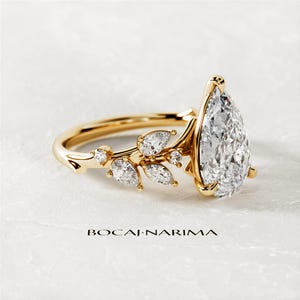 Puede incluir: Un anillo de compromiso de diamante de forma pera engastado en oro amarillo con un delicado dise&ntilde;o floral con diamantes m&aacute;s peque&ntilde;os. El anillo est&aacute; sobre un fondo blanco.