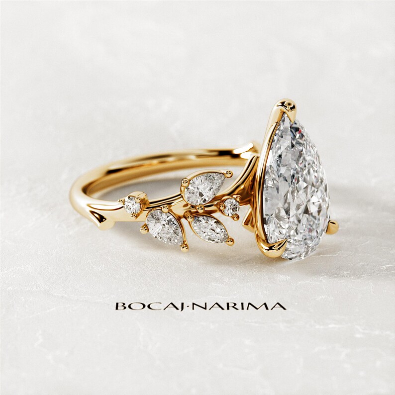 Puede incluir: Un anillo de compromiso de diamante de forma pera engastado en oro amarillo con un delicado dise&ntilde;o floral con diamantes m&aacute;s peque&ntilde;os. El anillo est&aacute; sobre un fondo blanco.
