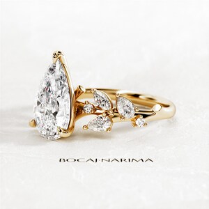Puede incluir: Un anillo de compromiso de oro con un diamante en forma de pera y un delicado dise&ntilde;o floral con diamantes m&aacute;s peque&ntilde;os. El anillo est&aacute; sobre un fondo blanco.