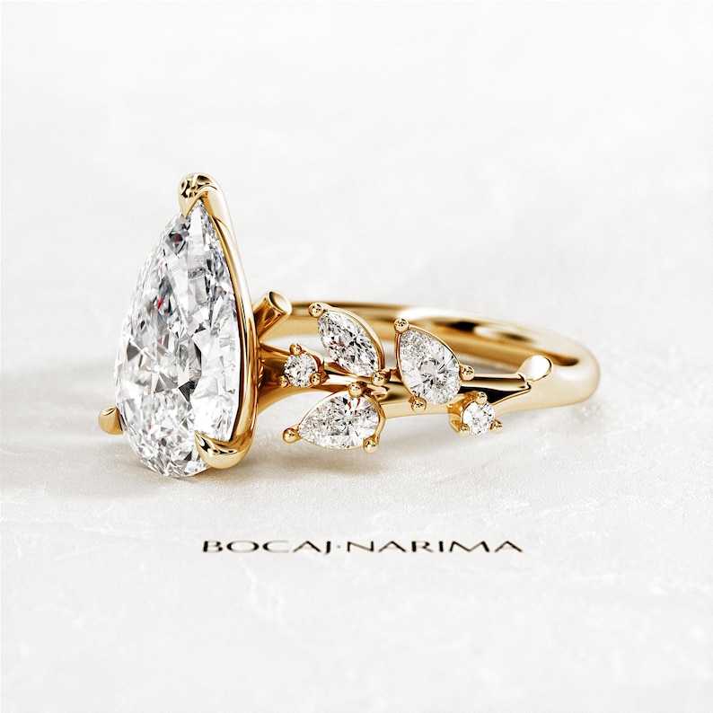 Puede incluir: Un anillo de compromiso de oro con un diamante en forma de pera y un delicado dise&ntilde;o floral con diamantes m&aacute;s peque&ntilde;os. El anillo est&aacute; sobre un fondo blanco.