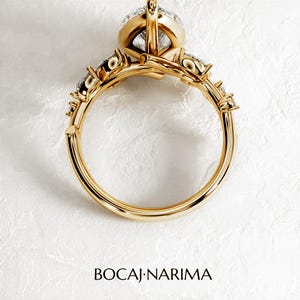 Puede incluir: Un anillo de oro con una piedra preciosa grande, redonda y transparente rodeada de piedras m&aacute;s peque&ntilde;as. El anillo est&aacute; sobre un fondo blanco.