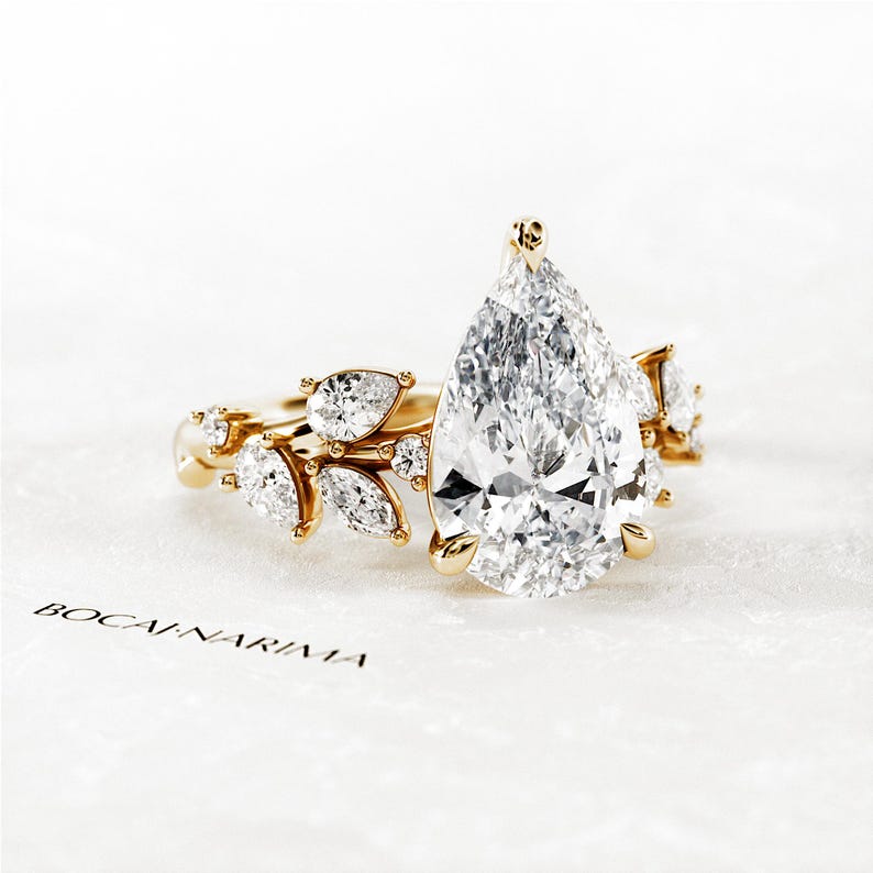 Puede incluir: Un anillo de compromiso de diamante de forma de pera engastado en oro con un delicado dise&ntilde;o floral con diamantes m&aacute;s peque&ntilde;os. El anillo se muestra sobre una superficie blanca con el nombre de la marca "BOCANARAMA" impreso en &eacute;l.