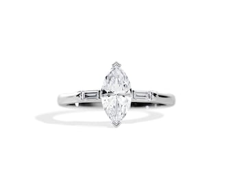 Puede incluir: Un anillo de compromiso de plata con un diamante de forma marquesa y dos diamantes más pequeños de forma baguette en la banda.