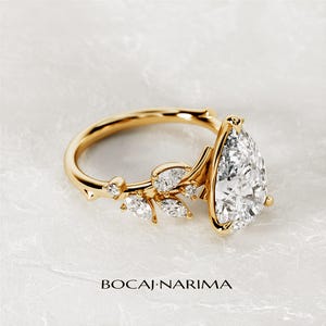 Puede incluir: Un anillo de compromiso de oro en forma de pera con un delicado dise&ntilde;o floral. El anillo presenta un gran diamante en forma de pera en el centro y diamantes m&aacute;s peque&ntilde;os engastados en un patr&oacute;n floral en la banda. El anillo est&aacute; colocado sobre un fondo blanco.