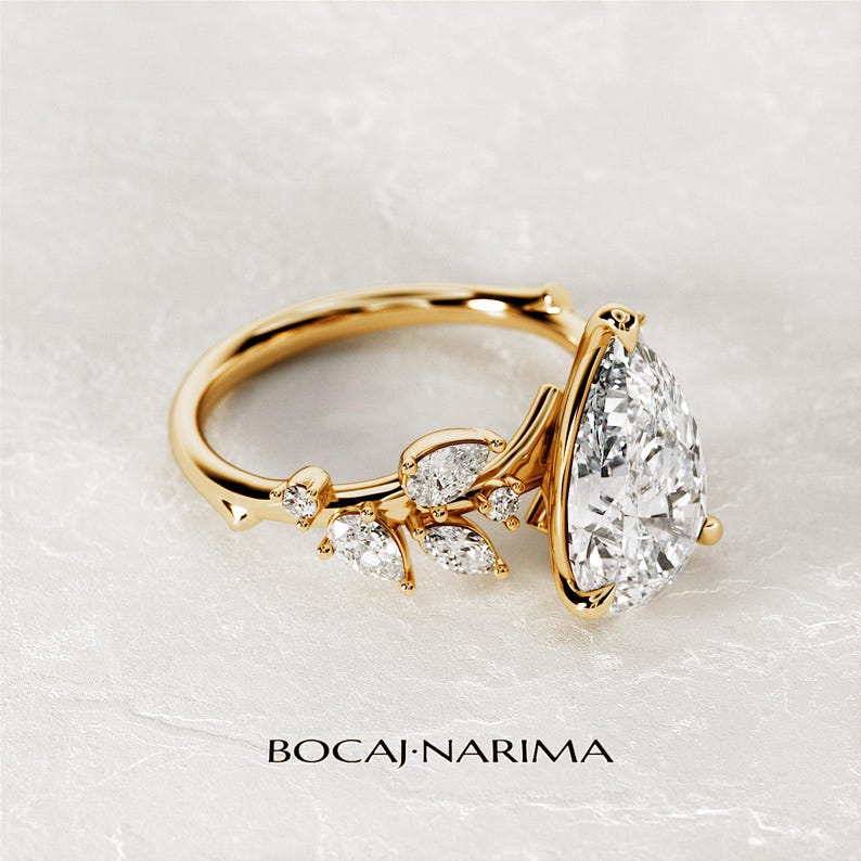 Puede incluir: Un anillo de compromiso de oro en forma de pera con un delicado dise&ntilde;o floral. El anillo presenta un gran diamante en forma de pera en el centro y diamantes m&aacute;s peque&ntilde;os engastados en un patr&oacute;n floral en la banda. El anillo est&aacute; colocado sobre un fondo blanco.