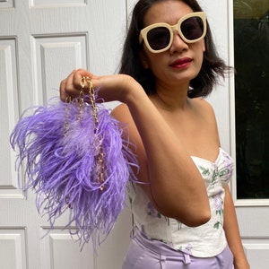 Pode incluir: Uma bolsa de penas roxa com uma alça de corrente dourada. A bolsa está sendo segurada por uma pessoa que está vestindo um top de espartilho branco floral e calças lavanda. A pessoa também está usando óculos de sol brancos.