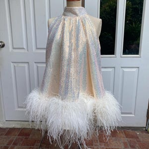Könnte beinhalten: Ein cremefarbenes Neckholder-Kleid, das mit schillernden Pailletten bedeckt ist. Das Kleid hat einen hohen Ausschnitt und ist mit einem flauschigen weißen Federbesatz versehen. Das Kleid wird auf einer Schaufensterpuppe präsentiert.