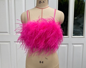 Peut inclure: Un haut court rose vif orné d'une couche dense de longues plumes légères recouvrant tout le buste. Le vêtement est conçu avec de fines bretelles roses assorties et une encolure droite, présenté sur un mannequin de couture beige.