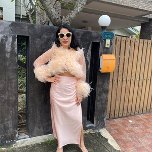 May include: A woman wearing a pink feather-trimmed top and a matching pink skirt. She is wearing silver shoes and sunglasses. The woman is standing in front of a wooden gate with a mailbox.