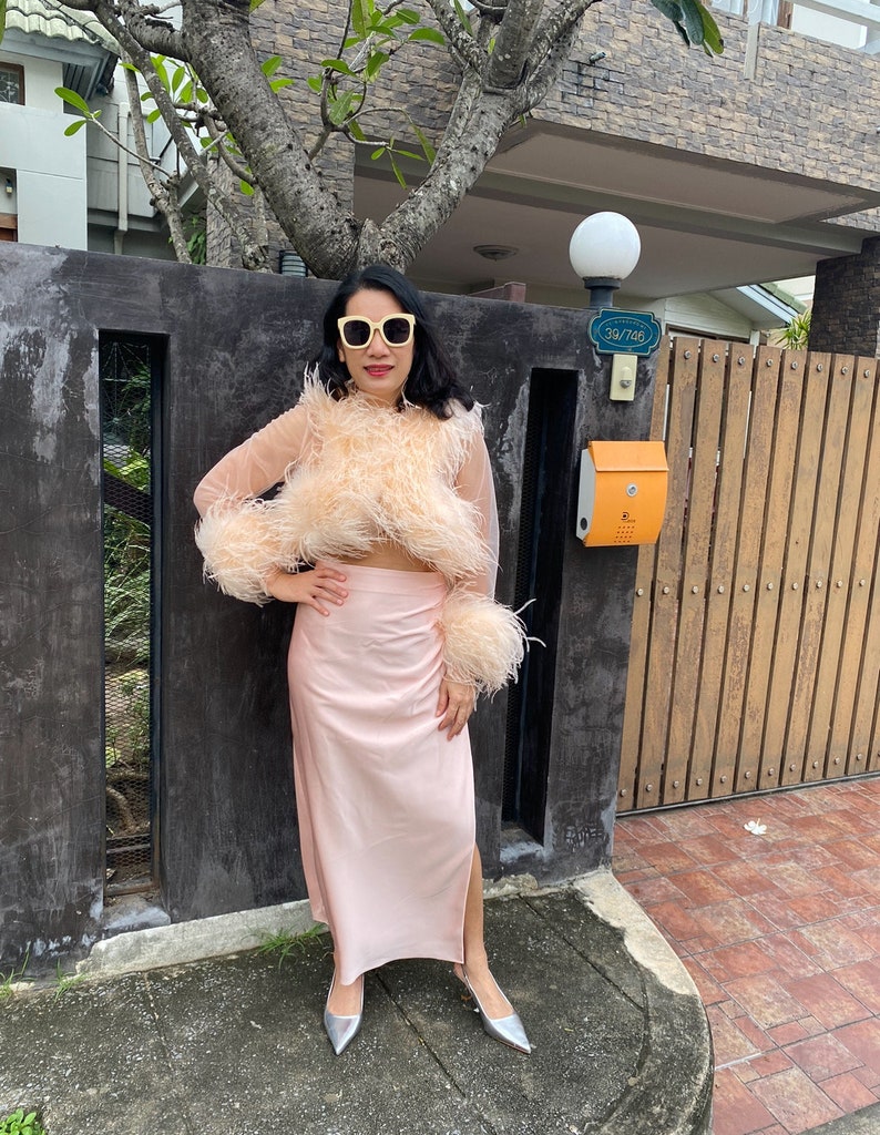 May include: A woman wearing a pink feather-trimmed top and a matching pink skirt. She is wearing silver shoes and sunglasses. The woman is standing in front of a wooden gate with a mailbox.