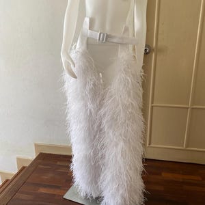 May include: A white, feathery skirt and belt set displayed on a mannequin. The skirt is made of long, soft white feathers, and the belt is white with a silver-colored buckle. The mannequin is positioned on a wooden surface.