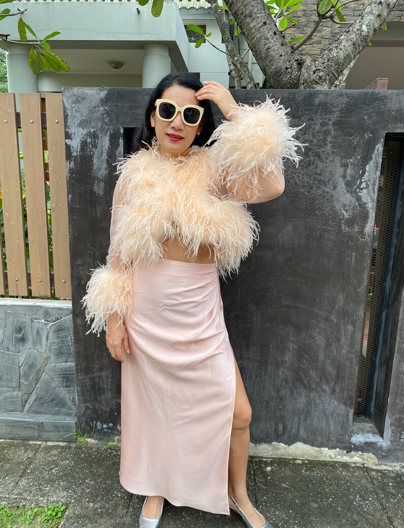 May include: A woman wearing a pink feather-trimmed top and a matching pink satin skirt with a slit. She is wearing white sunglasses and has her hair pulled back.
