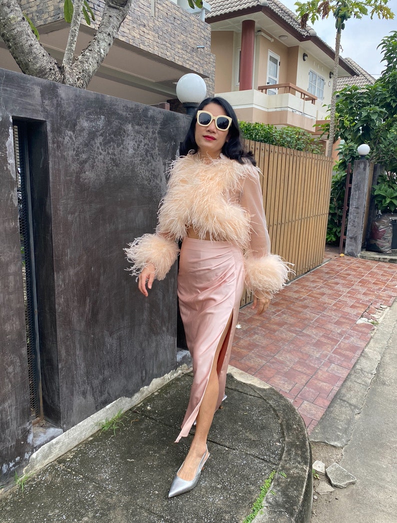 May include: A woman wearing a pink feather-trimmed top and a matching pink satin skirt with a side slit. She is wearing silver shoes and sunglasses.