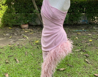 May include: A light pink, one-shoulder dress with a draped bodice and a feathered asymmetrical hemline. The dress is displayed on a mannequin, showcasing its elegant design and texture. The dress is made of a soft, flowing fabric.
