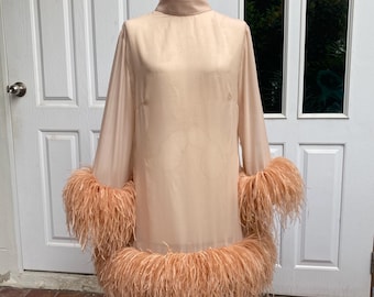 May include: A peach-coloured dress with long sleeves and a high neckline. The cuffs and hem are trimmed with fluffy, peach-coloured feathers. The dress is displayed on a mannequin in front of a white door.
