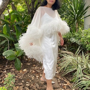 Pode incluir: Um vestido tipo slip dress branco de comprimento midi com uma capa transparente e fluida sobreposta. A capa é adornada com um acabamento espesso e volumoso de penas brancas ao longo da bainha, criando um efeito em camadas sobre o tecido.