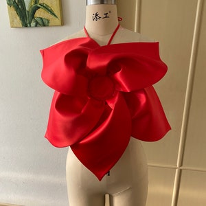 Könnte beinhalten: Ein rotes Satin-Haltertop mit einer großen Schleife vorne. Die Schleife ist zu einem Knoten gebunden und hat ein kreisförmiges Mittelstück. Das Oberteil befindet sich auf einer Schaufensterpuppe.