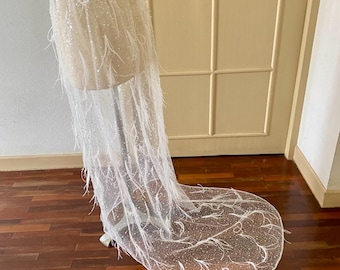 May include: A white, sheer skirt with a long train, adorned with delicate white feathers and sparkling embellishments. The skirt is displayed on a mannequin, showcasing its flowing design. The skirt is made of a lightweight fabric.