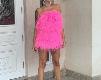 May include: A pink strapless mini dress made of feathers. The dress is short and has a fluffy texture. The model is wearing the dress with silver jewelry and heels.