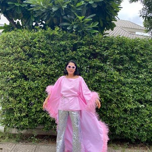 May include: A woman wearing a pink top with a long feathered train and silver sequined trousers. She is standing in front of a green hedge.