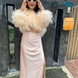 May include: A woman wearing a pink feather crop top and a matching pink satin skirt with a slit. She is wearing yellow sunglasses and silver heels. The woman is standing in front of a black gate with a mailbox and a sign that reads "39/746".