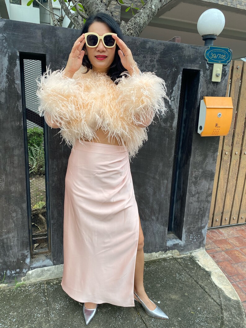 May include: A woman wearing a pink feather crop top and a matching pink satin skirt with a slit. She is wearing yellow sunglasses and silver heels. The woman is standing in front of a black gate with a mailbox and a sign that reads "39/746".