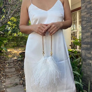 Könnte beinhalten: Weißes Satin-Slip-Kleid mit Spaghettiträgern und V-Ausschnitt. Eine kleine, weiße, federartige Clutch mit goldener Kette wird vor dem Kleid gehalten. Das Kleid ist knielang.