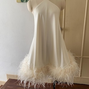 Op de afbeelding: Een crèmekleurige halterjurk met een uitlopende rok en een rand van pluizige veren aan de zoom. De jurk wordt getoond op een mannequin, die het elegante ontwerp en de vloeiende silhouet laat zien. Het is een formeel kledingstuk.