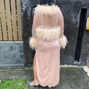 May include: A long pink satin skirt with a slit up the side, paired with a sheer pink top with feather trim. The top has a low back and is worn over a nude bra.
