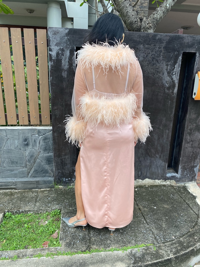 May include: A long pink satin skirt with a slit up the side, paired with a sheer pink top with feather trim. The top has a low back and is worn over a nude bra.